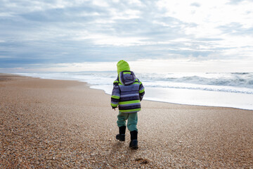 A small child in warm clothes walks along the seashore during the cold season. Activity and walks. Back view.