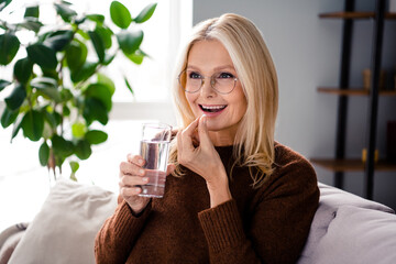 Photo of adorable cute woman pensioner sitting on comfy couch in living room hold glass drink pills...