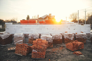 Building materials at construction site. Autoclaved aerated blocks on concrete foundation and bricks in evening light, process of house building. Stacks of white aerated blocks for laying.