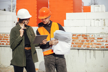 Stylish woman architect with tablet  and contractor man checking blueprints at construction site. Young engineer or construction workers in hardhat looking at plans of new modern house - obrazy, fototapety, plakaty