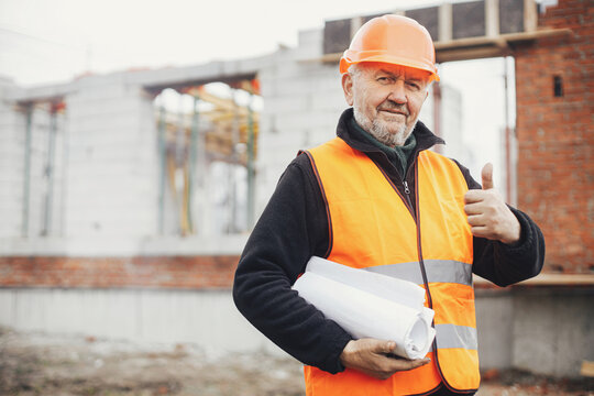 Senior male foreman in hardhat approving building new modern house according to blueprints. Portrait of happy man engineer or construction worker with plans at construction site