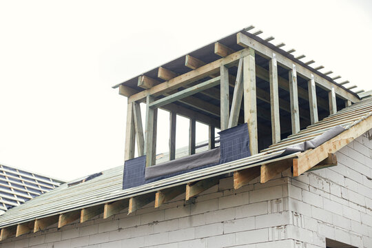 Unfinished house. Wooden roof framing with vapor barrier and dormer on block walls with windows. New modern farmhouse construction. Timber trusses Rafters and beams on aerated concrete blocks