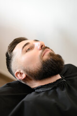 Obraz premium A young man with a beard in a black cape sits on a chair in a barbershop. The client is waiting for a haircut of the beard.