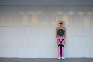 Attractive young gay man, heavily makeup, with pink hair, sunglasses, leather top and pants, posing leaning against a white stone block wall. LGTBIQ+ concept, gay, pride, makeup, fashion, trend.