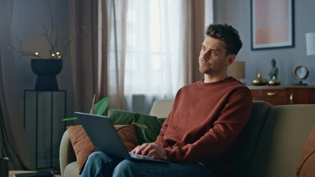 Concerned writer typing laptop on living room couch. Serious man feeling burnout