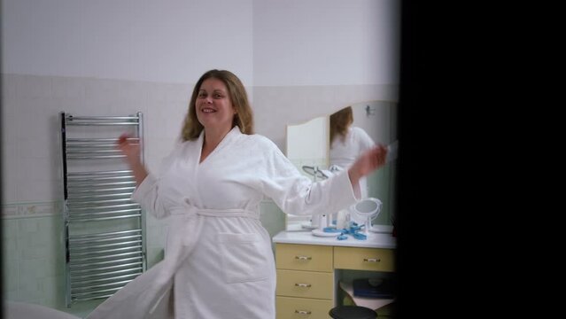 Slow motion. A woman enters the bathroom in a white bathrobe in a great mood. A woman is spinning with her arms raised in the middle of the bathroom and smilingly posing looking at herself in the mirr