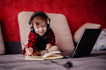 A young boy with headphones focuses intently on writing in a notebook, with a laptop open in front of him, embracing the joy of learning