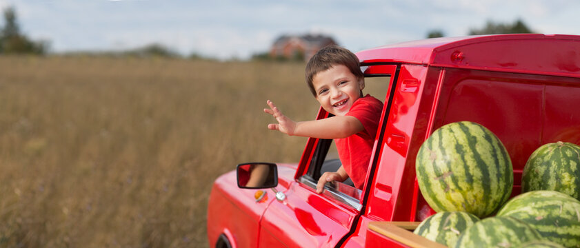 child boy waves from the cab of a red pickup truck  with watermelons.  banner. copy space.summer time in nature in the village