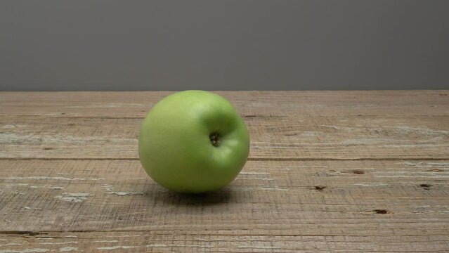 A green apple rolls on a wooden surface on a gray background.