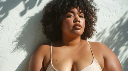 A woman with curly hair wearing a white tank top leaning against a white wall with sunlight casting shadows.