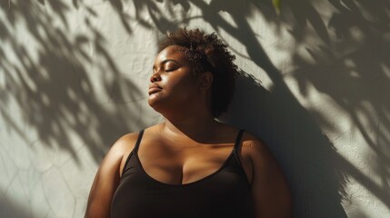 A woman with curly hair wearing a black tank top leaning against a wall with her eyes closed basking in the sunlight filtering through leaves.