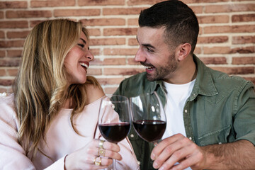 Joyful young adult couple toasting with wine sitting at a table. Laughing man and woman drinking with glasses at home.