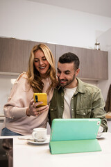 Happy joyful man and woman with smartphone and tablet having a coffee cup. Young adult couple reading together online device from home in a modern kitchen.
