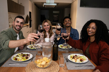 Smiling people toasting eating and drinking on a modern apartment. Diverse group of happy friends celebrating a diner at home looking at camera. 