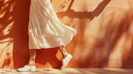 A person in a flowing white dress and white sneakers walking past a textured orange wall with sunlight casting shadows.