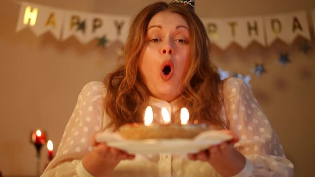 A woman in a birthday-cap holds a birthday cake with burning candles in her hands. A woman blows out the candles on a cake and looks at the cake smiling