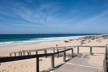 A tranquil beach scene in Comporta Beach, Setubal, Portugal, with a wooden boardwalk leading to a sandy beach against a backdrop of a clear blue sky and rolling waves