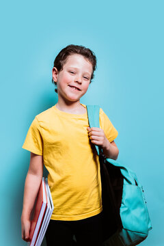 A young student in a yellow tee grins while holding textbooks and wearing a backpack, set against a light blue background