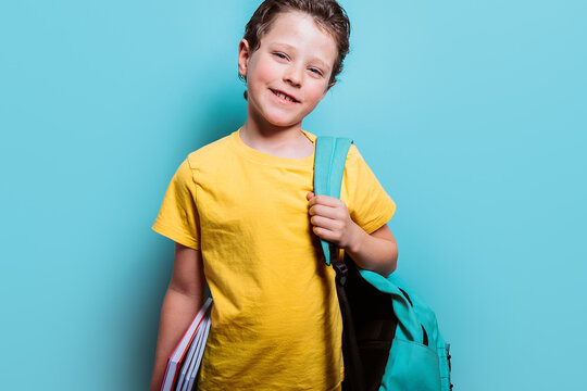 A young student in a yellow tee grins while holding textbooks and wearing a backpack, set against a light blue background
