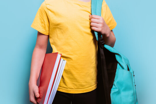 Anonymous young student wearing a yellow tee while holding school books and wearing a rucksack, against a light blue background