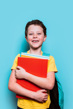 A smiling school boy in a yellow shirt tightly hugs his school books, with a backpack over one shoulder, against a blue background
