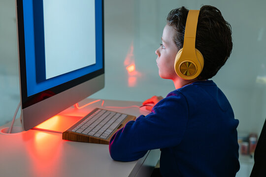 A child is intently using a computer, illuminated by the warm backlight of the monitor, with headphones on