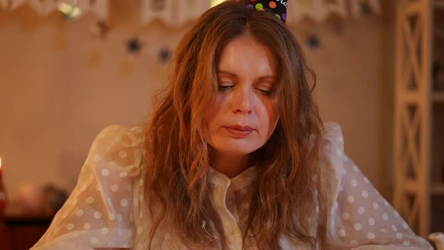 A woman in a birthday cap licks her fingers with the remains of a birthday cake on them and looks at the camera, while sitting at the table on her birthday