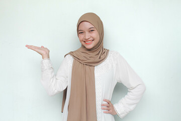 Young Asian Muslim woman wearing hijab, sweet smiling and see camera while pointing and showing to copy space beside her. Isolated in gray background.