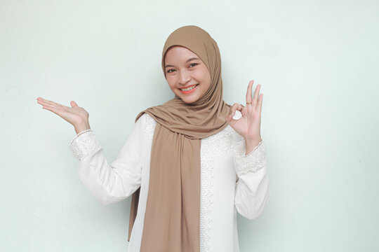 Young Asian Muslim woman wearing hijab, smiling and see camera while pointing and showing to copy space beside her with ok gesture. Isolated in gray background.