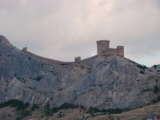 An ancient medieval fortress on the edge of a cliff is surrounded by large, bright clouds. Landscape of a tourist and historical place with a cold blue tint.