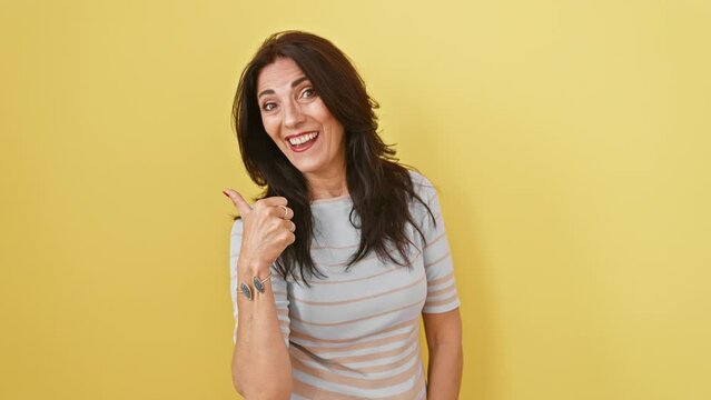 Joyful mature hispanic woman with open mouth, confidently standing, pointing thumb up, gesturing to side, on isolated yellow background.