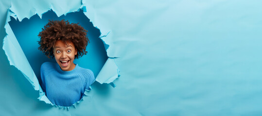 Cheerful amazed dark skinned Afro American woman indicates at blank space demonstrates advertisement wears blue jumper has happy expression breaks through paper background. Promotion concept - obrazy, fototapety, plakaty