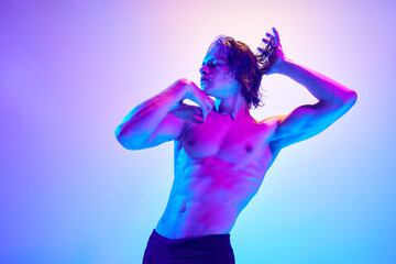 Portrait of athlete man with wet hair posing raising hands in vibrant neon light against gradient blue-pink background. Concept of natural beauty people, fitness, male health and wellness, masculinity
