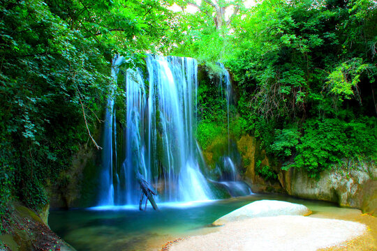 Long-exposure shot of Cascata "de lu Vagnat&ograve;" in Sarnano with fluent streams of water coming down, washing the mossy side behind them, gathering into a basin below between white boulders and greenery