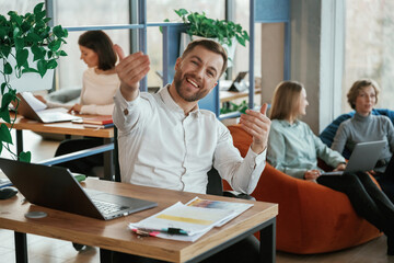 Happy man is smiling, sitting by table. People are working in the office with bean bags chairs in it