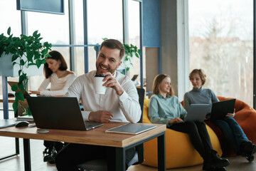 Man is smiling, sitting by the table with laptop. People are working in the office with bean bags chairs in it