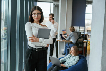 Beautiful woman is standing and holding laptop. People are working in the office with bean bags chairs in it