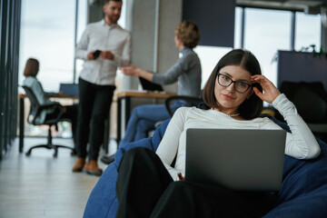 Woman in glasses is sitting with laptop. People are working in the office with bean bags chairs in it