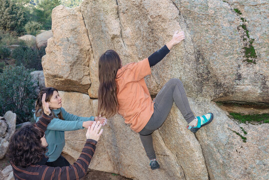 A climber reaches for a high hold, supported by attentive teammates, amidst a rugged granite boulder landscape