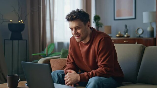 Smiling man video calling by laptop at apartment couch. Guy have online meeting