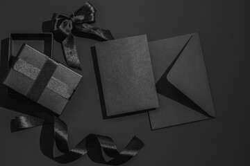 Black gift box with satin ribbons and bow and a black envelope on a black background. Greeting card.