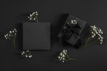 Black gift box and black card on a black background with space for text and sprigs of gypsophila....