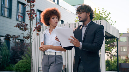 Indian Arabian male man businessman holding papers documents corporate discuss result plans with African American woman colleague businesswoman walking downtown in city multiracial business people - Powered by Adobe