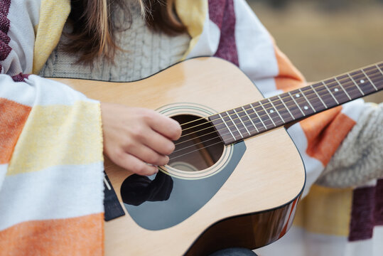 Close up of a girl playing guitar outdoors. Young girl covered in warm plaid playing acoustic guitar in autumn park