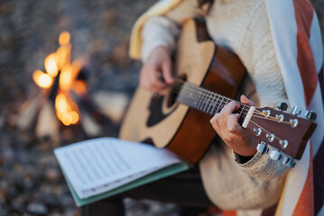 Close up of a girl guitarist practicing on nature near the camp fire. Warm evening shot of a girl...