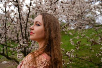beautiful blond natural hair woman portrait in pink outfit is posing in botanical garden park near white blooming tree with flowers. Spring and purity, natural beauty. 
