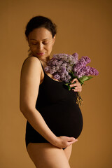Delightful pregnant woman in black bodysuit, posing with purple lilacs on beige background,...