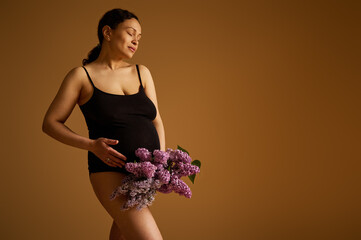 Authentic pregnant woman in black bodysuit, smiles while strokes her belly in last trimester of...