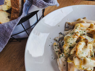Meal in the restaurant. fresh and delicious penne pasta sprinkled with parmesan and spices. next to it is a napkin and a basket of fragrant freshly baked bread. Food is on the wooden table