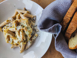 Meal in the restaurant. fresh and delicious penne pasta sprinkled with parmesan and spices. next to it is a napkin and a basket of fragrant freshly baked bread. Food is on the wooden table